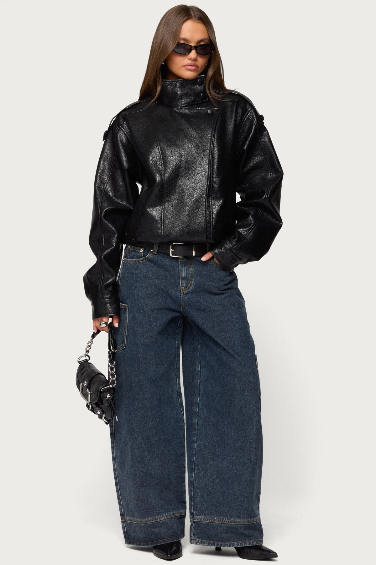 Full-length photo of a woman with sunglasses in a black oversized faux leather jacket, wide dark cargo jeans, belt and a small black chain bag