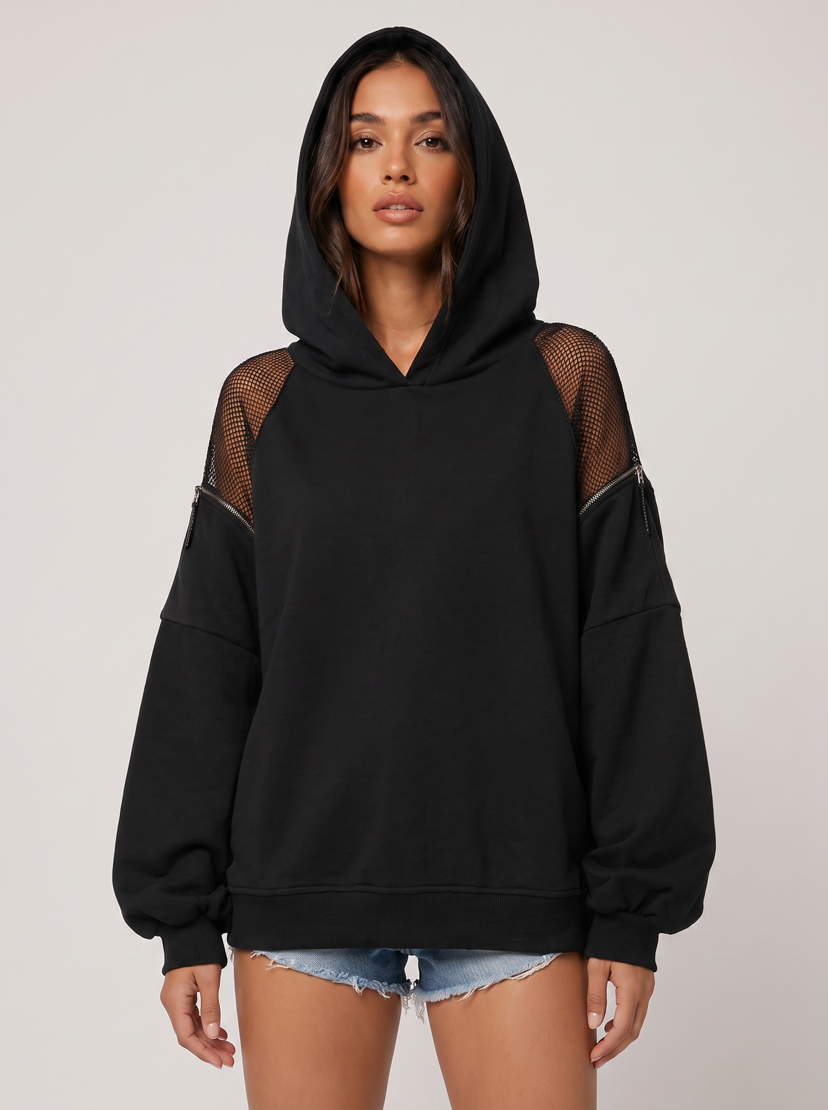 Full-length photo of a woman in a black oversized hoodie with mesh shoulder details and light denim shorts