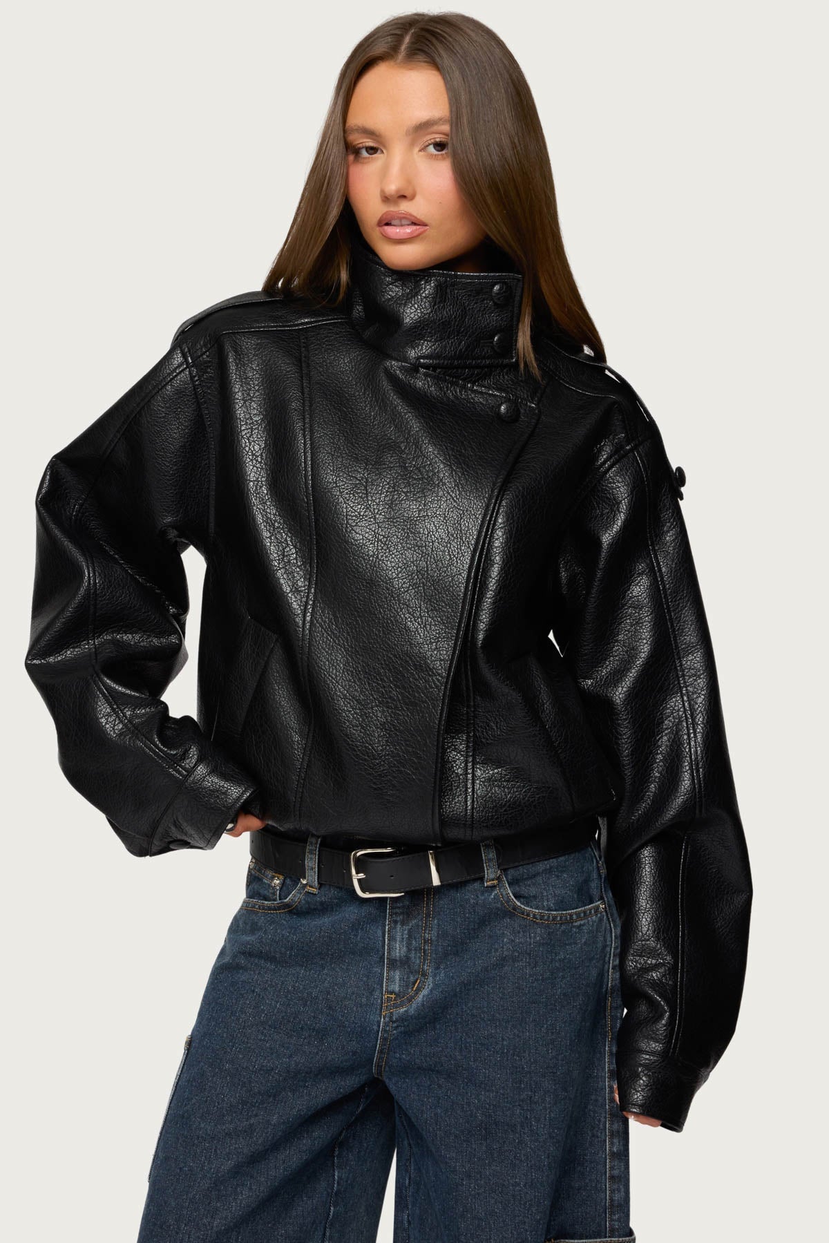 Woman in a black oversized faux leather jacket with a high stand-up collar and asymmetric snap placket, with dark baggy jeans